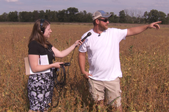 A reporter stands with an interviewee in a field, where they're discussing farm conservation.
